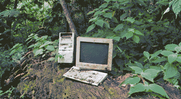 A dirty desktop computer tower, monitor, and keyboard on the ground in a forest.