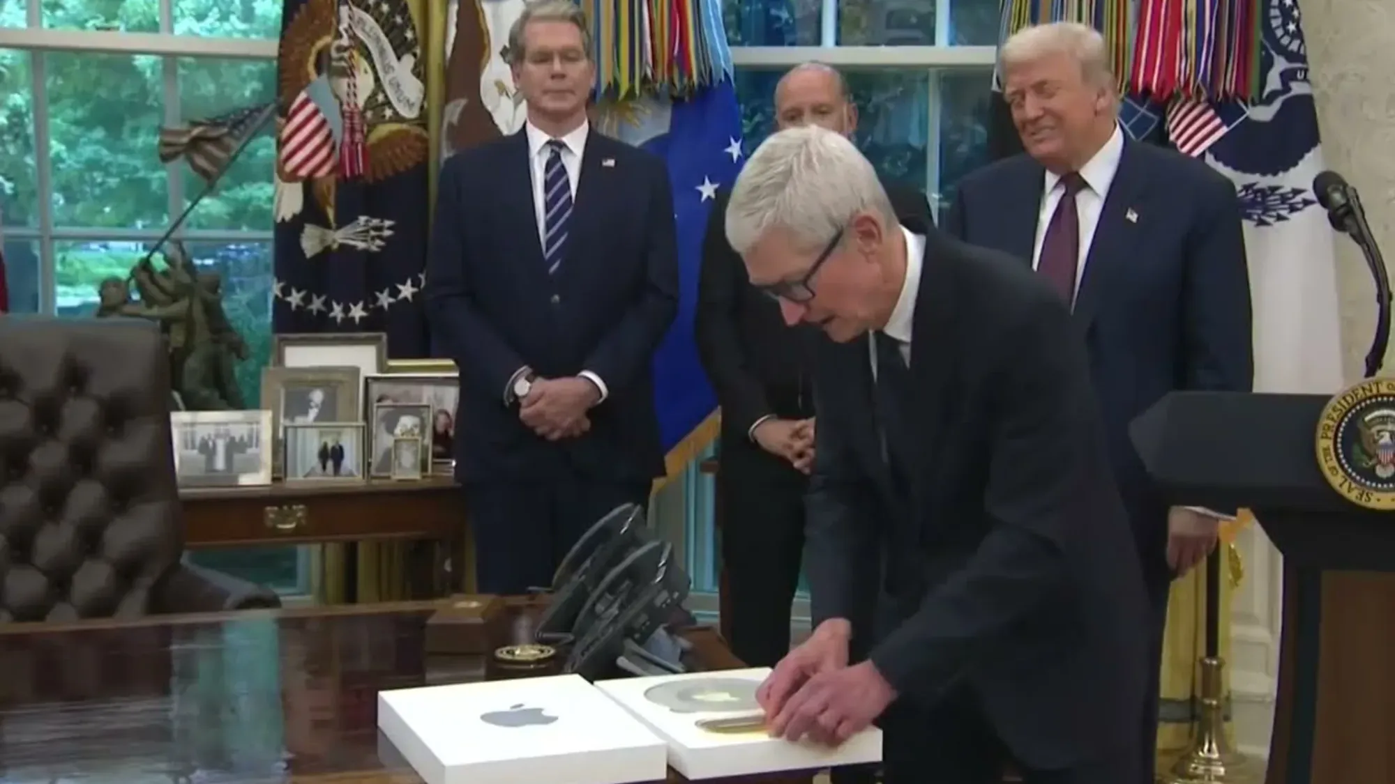 Apple CEO Tim Cook opens a white Apple-branded box on the Resolute Desk in the Oval Office, August 2025. President Trump watches from behind. Cook is presenting a custom glass-and-gold plaque commemorating Apple's “American Manufacturing Program” — part of a US$100 billion domestic investment pledge that secured tariff relief for Apple’s Chinese supply chain. 6 August 2025.