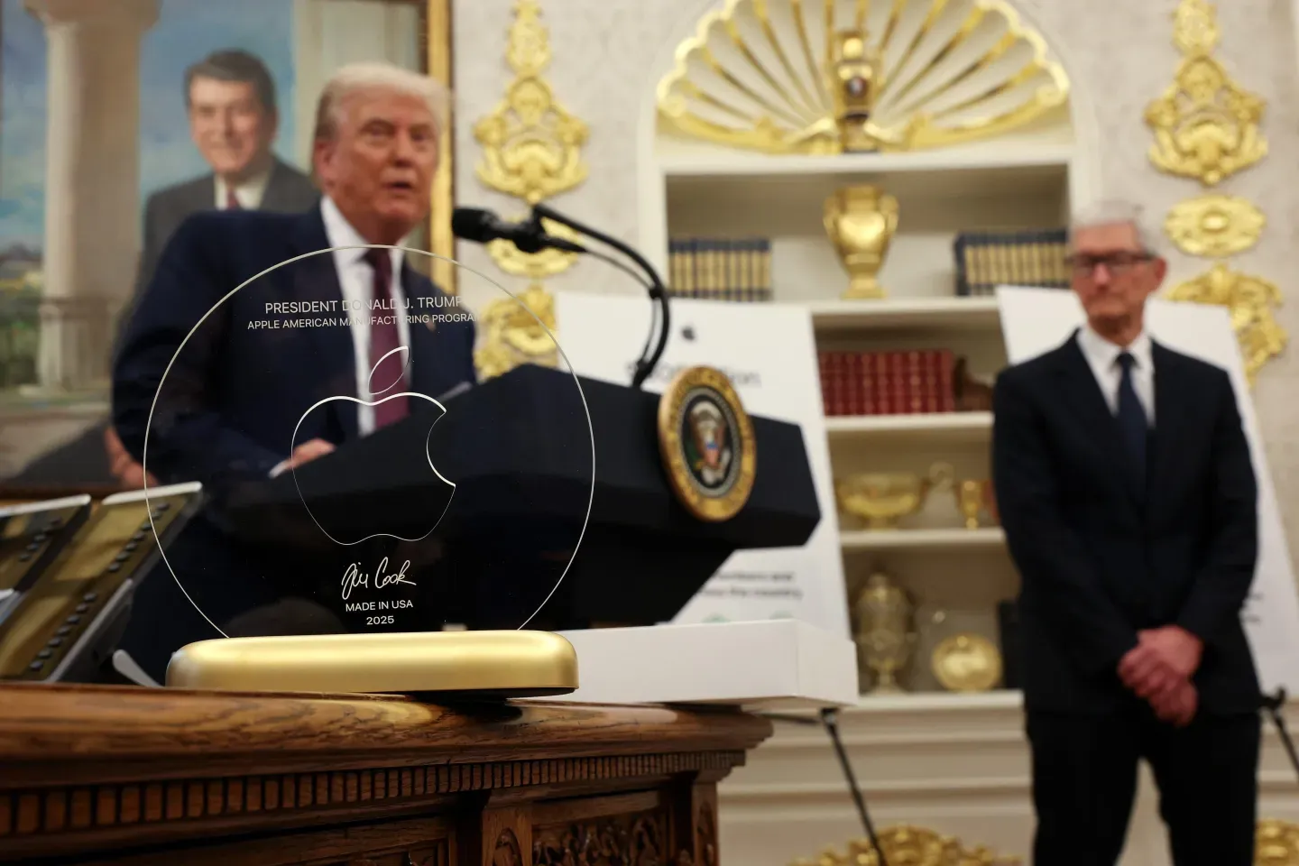 The gift: a glass disc etched with the Apple logo, inscribed “President Donald J. Trump — Apple American Manufacturing Program,” signed by Cook, “Made in USA, 2025,” mounted on a 24-karat gold base. Behind it, Trump stands beneath a portrait of Ronald Reagan, with Apple CEO Tim Cook to his left. The glass was manufactured by Corning in Kentucky; the gold base was sourced from Utah. 6 August 2025.